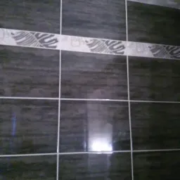 Ciemne, błyszczące płytki ścienne z szarym wzorem ułożone w regularną siatkę z białymi fugami, fragment ściany łazienki.