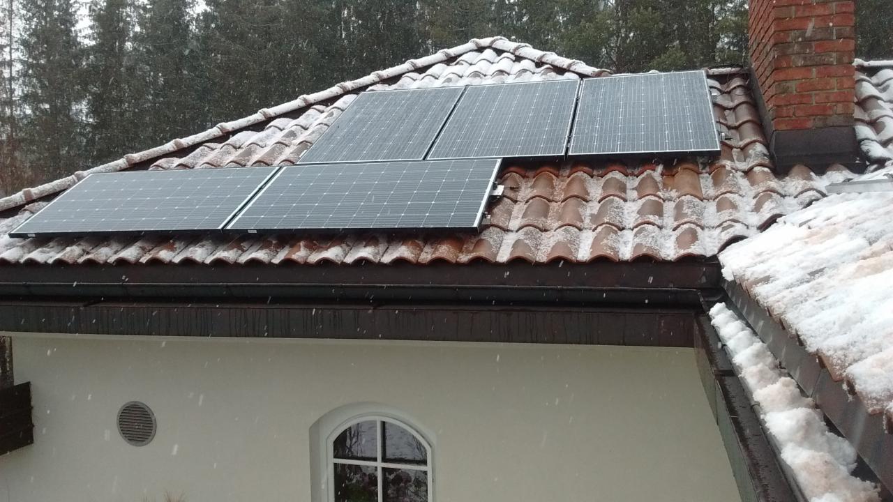 Panele fotowoltaiczne na dachu pokrytym śniegiem, widoczne z dołu podczas opadów śniegu. Dachówka w kolorze ceglastym, komin z czerwonej cegły.