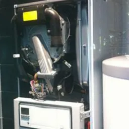 Otwarty kocioł grzewczy z widocznymi elementami wewnętrznymi, w tym palnikiem i okablowaniem, obok biały zasobnik ciepłej wody.