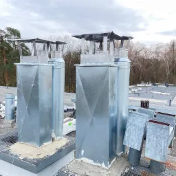 Nowe, metalowe kominy wentylacyjne z daszkami na płaskim dachu, świeżo osadzone w zaprawie, z widocznymi śladami prac montażowych i narzędziami w tle, na tle drzew i pochmurnego nieba.