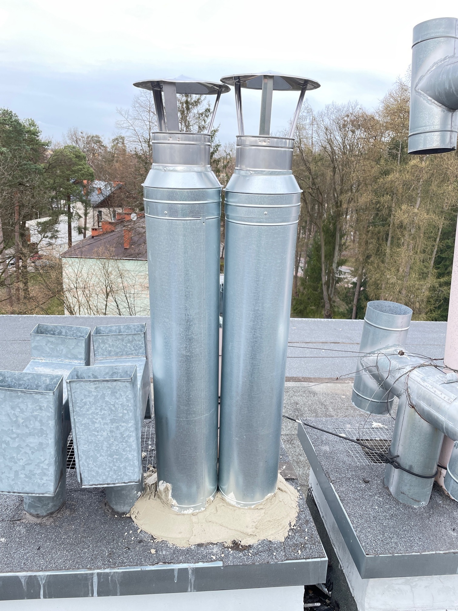 Dwa metalowe kominy wentylacyjne z daszkami na płaskim dachu, z widocznymi elementami instalacji i uszczelnieniami, na tle drzew i zabudowań.