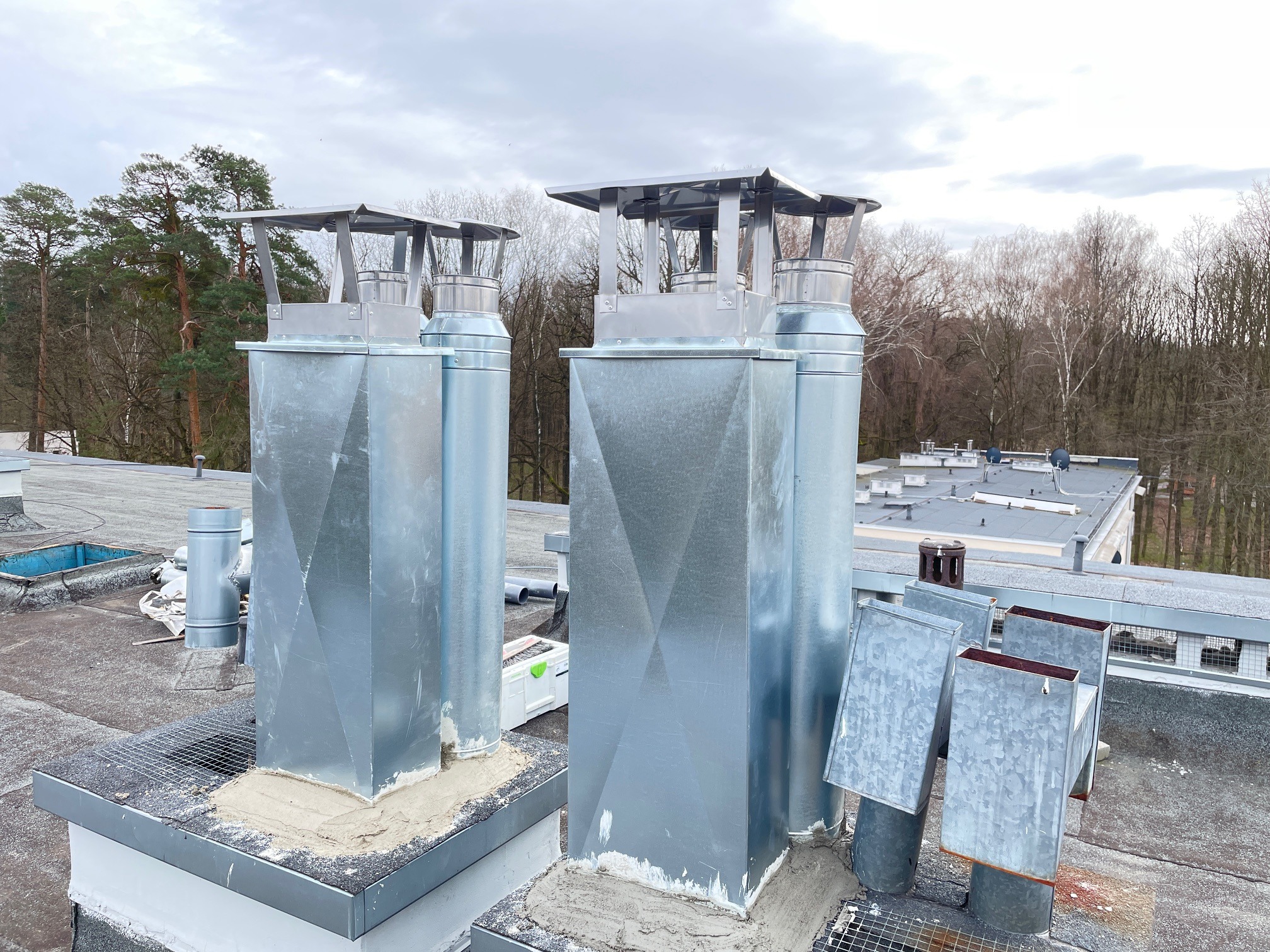Nowe, metalowe kominy wentylacyjne z daszkami na płaskim dachu, świeżo osadzone w zaprawie, z widocznymi śladami prac montażowych i narzędziami w tle, na tle drzew i pochmurnego nieba.