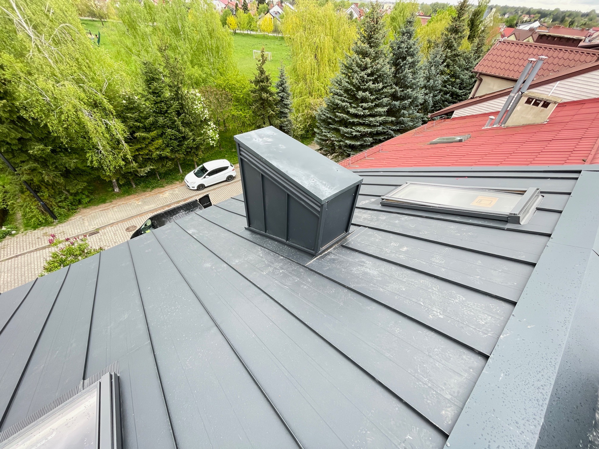 Szary dach z blachy na rąbek stojący, z lukarnami i kominem wentylacyjnym, widok z góry na tle zieleni i sąsiednich domów z czerwoną dachówką.