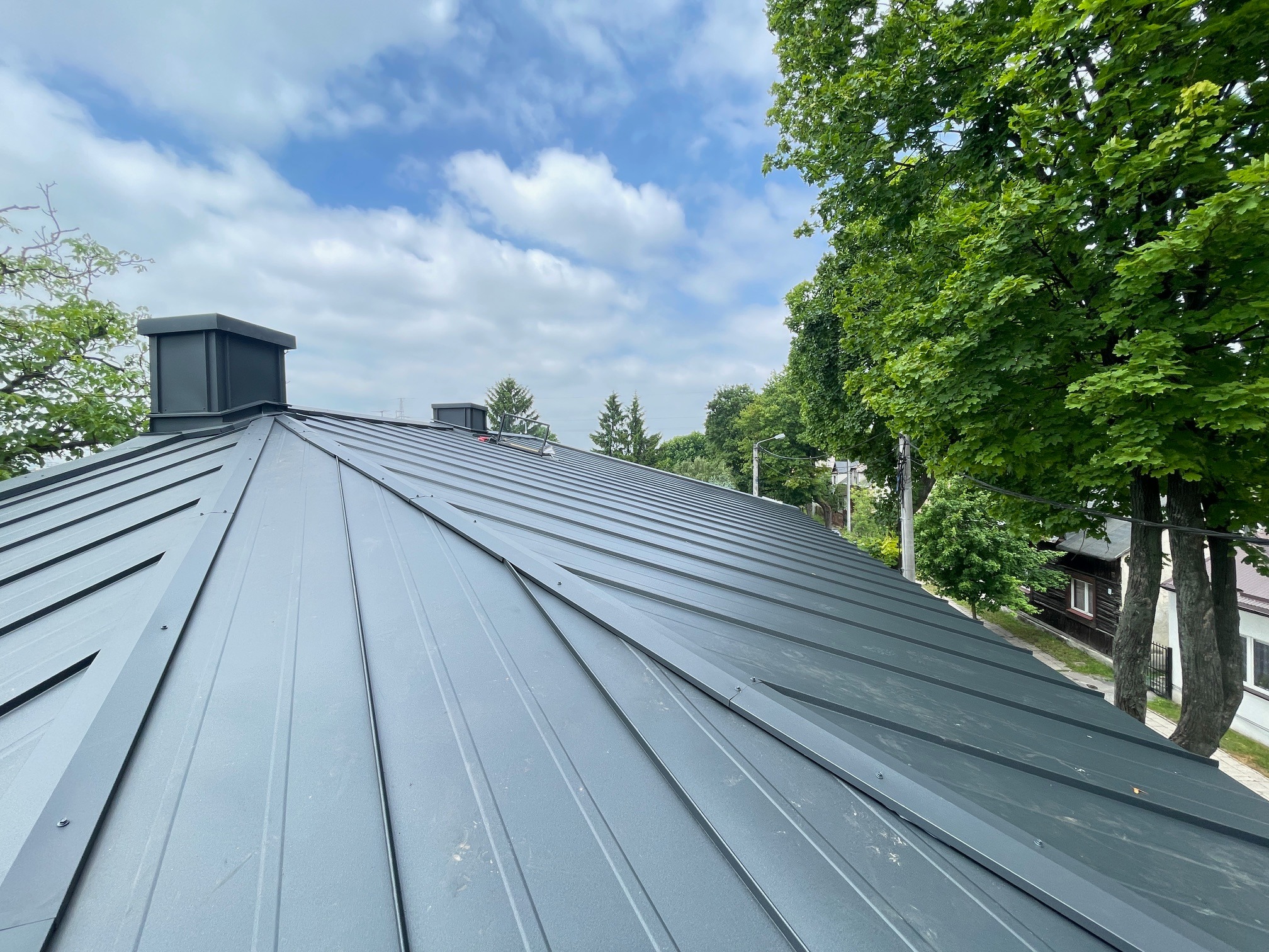 Nowoczesny dach z blachy na rąbek stojący w kolorze grafitowym, widok z góry, z kominem i elementami wentylacyjnymi, na tle błękitnego nieba z chmurami i zieleni drzew.