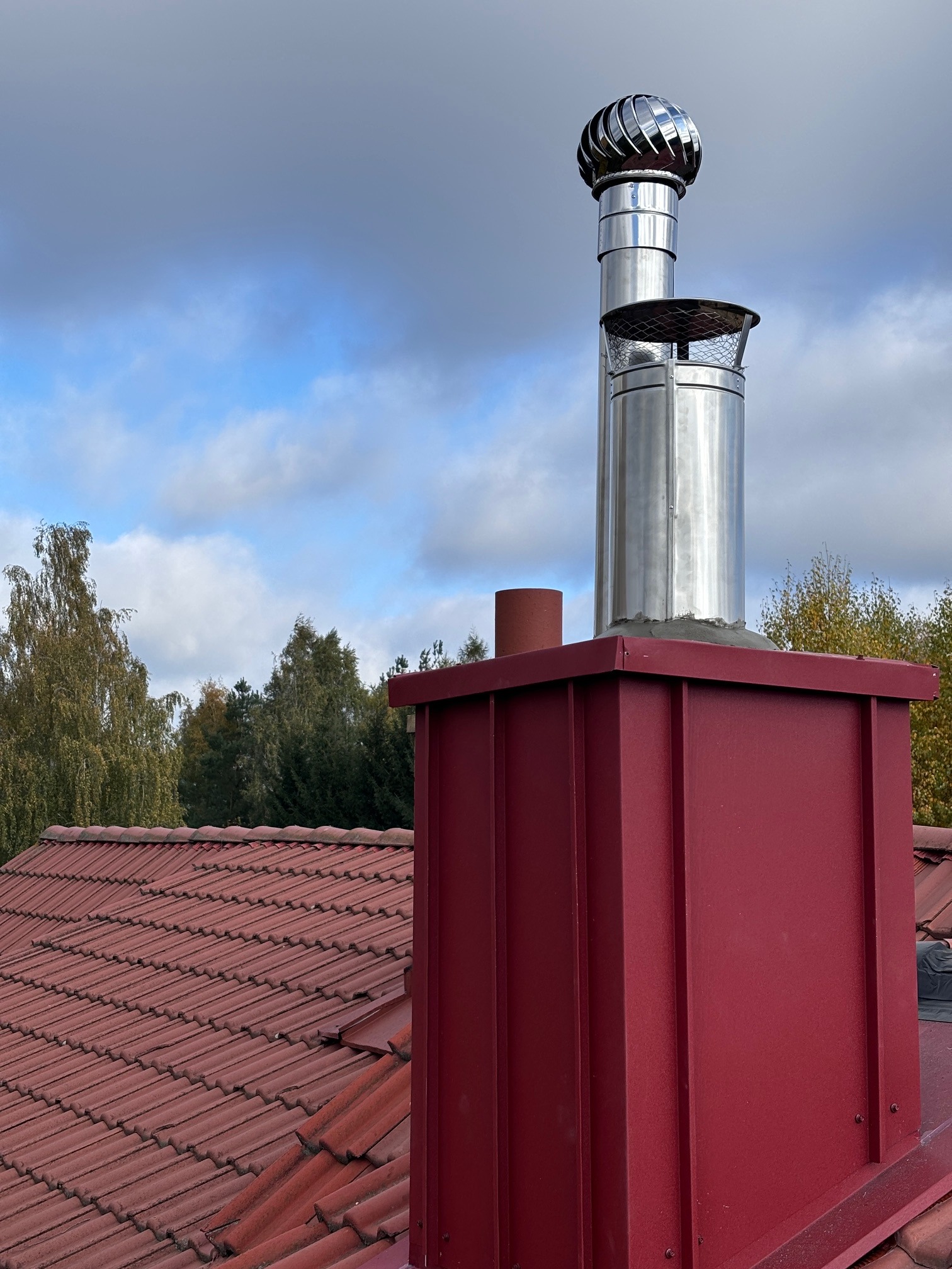 Czerwona obudowa komina z nierdzewną nasadą obrotową na tle błękitnego nieba z chmurami i fragmentu dachu z czerwonej dachówki ceramicznej.