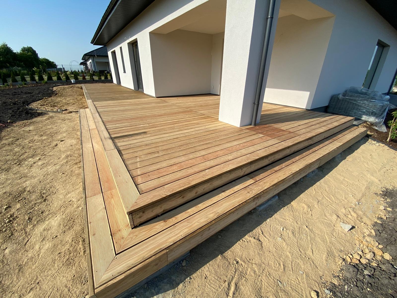 Nowoczesny taras z jasnego drewna z trzema stopniami, przylegający do białego domu z kolumną, widoczne przygotowane podłoże i elementy wyposażenia w folii.