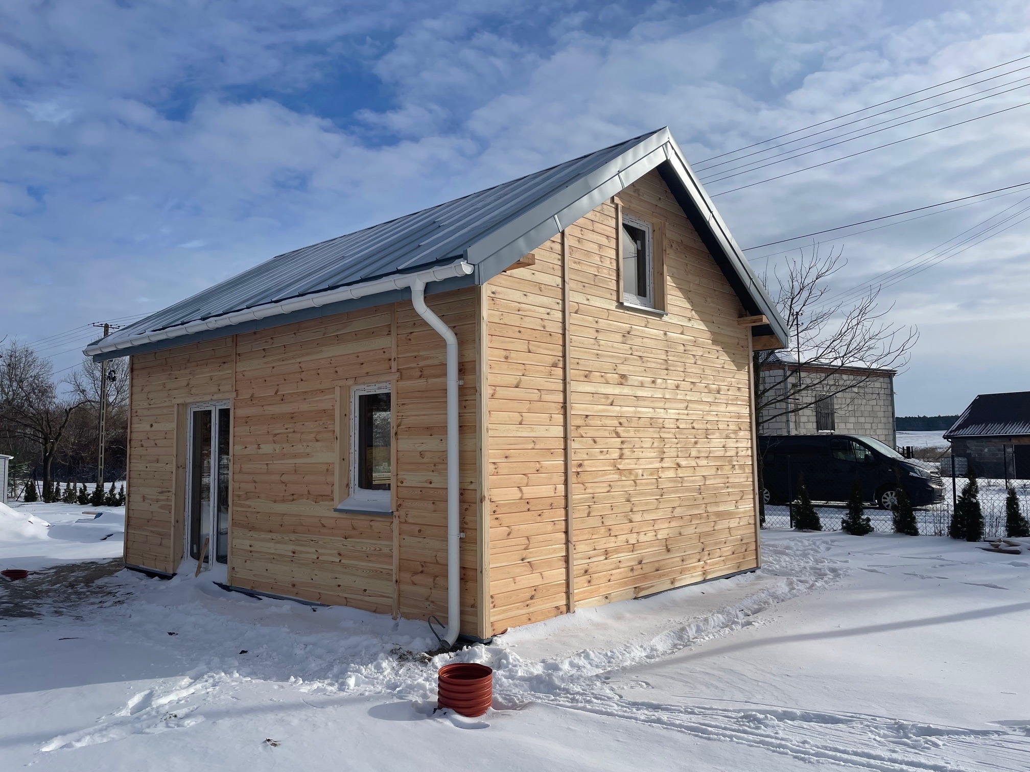 Surowy, drewniany dom szkieletowy z metalowym dachem, częściowo ośnieżony, z widoczną stolarką okienną i drzwiową, stojący na tle zimowego krajobrazu z niebieskim niebem i chmurami.