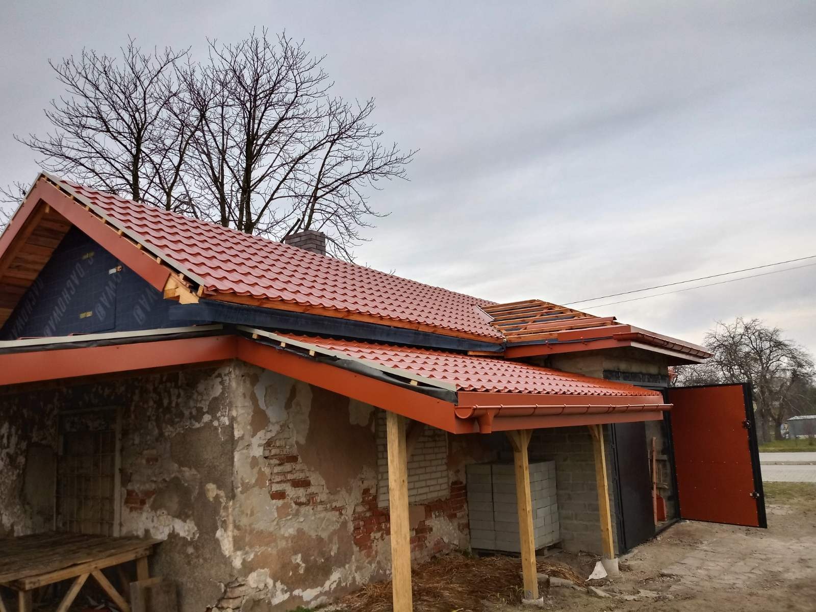 Ceglastoczerwony dach z blachy trapezowej na budynku w trakcie remontu, widoczne elementy konstrukcji dachu i rynny w kolorze dopasowanym do dachu, fragmenty elewacji z odsłoniętą cegłą i resztkami...