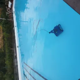 Niebieski automatyczny odkurzacz basenowy z przewodem pływający w czystej wodzie basenu zewnętrznego, widok z góry, drzewa w tle.