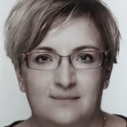 Portret kobiety z krótkimi, jasnymi włosami, w okularach z cienkimi, czerwonymi oprawkami, z delikatnym uśmiechem i srebrną biżuterią.