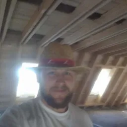 Mężczyzna w słomkowym kapeluszu z czerwoną wstążką na tle drewnianej konstrukcji dachu w trakcie budowy, widoczne elementy więźby dachowej i otwory okienne.