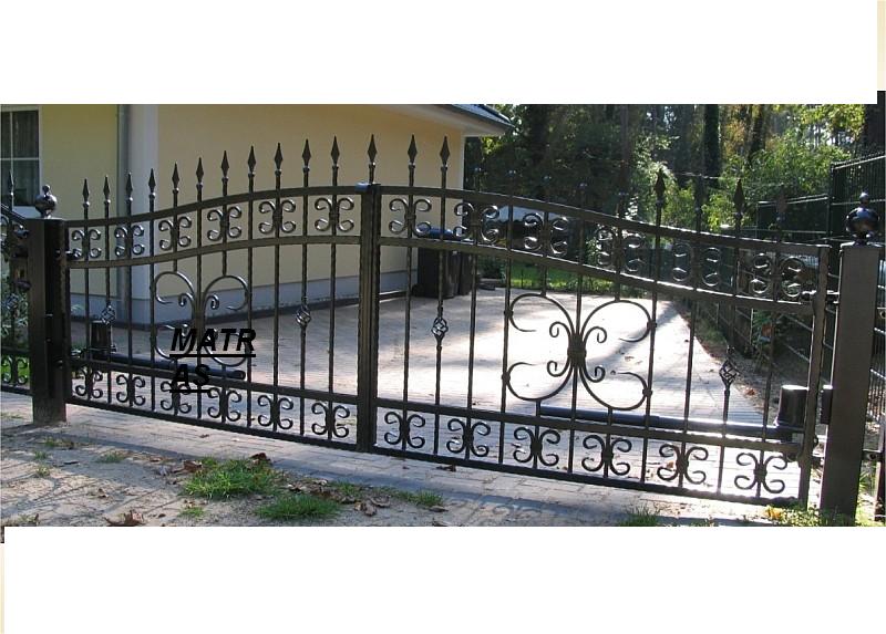 Kuta, metalowa brama przesuwna z dekoracyjnymi elementami, widoczny automat do bramy, na tle domu i drzew.