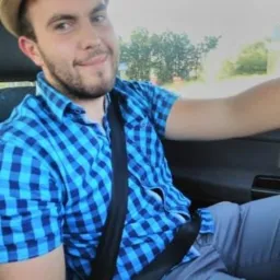 Uśmiechnięty mężczyzna w kapeluszu i niebieskiej koszuli w kratę, siedzący za kierownicą samochodu z zapiętym pasem bezpieczeństwa. Widok z perspektywy pasażera.