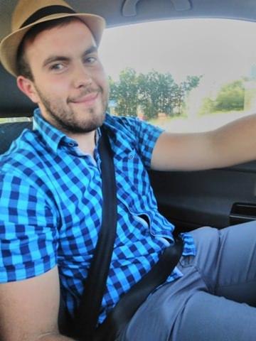 Uśmiechnięty mężczyzna w kapeluszu i niebieskiej koszuli w kratę, siedzący za kierownicą samochodu z zapiętym pasem bezpieczeństwa. Widok z perspektywy pasażera.