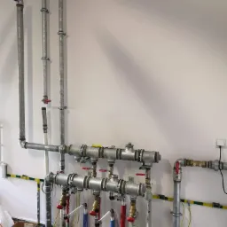 Szara instalacja rurowa z zaworami i izolacją na ścianie, widoczne elementy montażowe i przewody elektryczne.