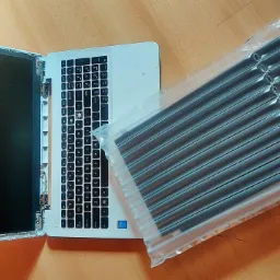 Otwarty laptop z odłączonym ekranem, obok opakowanie ochronne z czarnymi tubami powietrznymi, widoczne ślady demontażu obudowy.