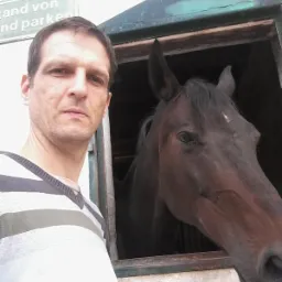 Mężczyzna w paski pozuje z koniem w stajni, widoczne tablice informacyjne. Nietypowe ujęcie.