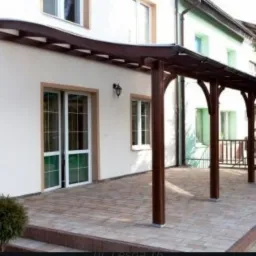 Zadaszony taras z drewnianą konstrukcją wspartą na łukowatych słupach, przylegający do białego budynku z jasnymi oknami i drzwiami, wyłożony kostką brukową.