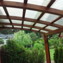 Zadaszenie z ciemnego drewna i przezroczystego falistego tworzywa, widok od wewnątrz, w tle zieleń ogrodu.