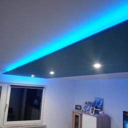 Dan-Bud - Podwieszany sufit z niebieskim oświetleniem LED i punktowymi lampami w pokoju, widok na okno i meble.