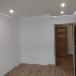 Dan-Bud - Pusty pokój po remoncie z drewnianą podłogą, świeżo pomalowanymi białymi ścianami, podwieszanym sufitem z oświetleniem LED i otwartymi drewnianymi drzwiami w tle.