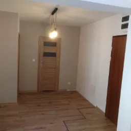 Korytarz po remoncie z drewnopodobną podłogą, dwiema drewnianymi ościeżnicami i drzwiami, oraz nowoczesną lampą sufitową.