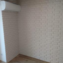 Dan-Bud - Ściana wykończona dekoracyjną, białą cegłą, z zamontowanym klimatyzatorem i fragmentem drewnianej podłogi.