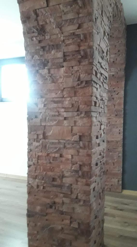 Aranżacja wnętrza z kolumną i łukiem wykończonymi dekoracyjną okładziną z piaskowca w odcieniach brązu i beżu, na podłodze panele imitujące drewno.