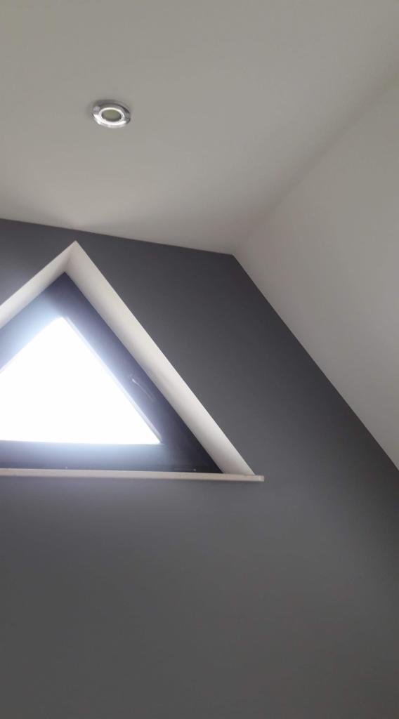 Szara ściana w pokoju na poddaszu z trójkątnym oknem i białym sufitem z wbudowanym oświetleniem.