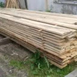 Stos surowej, jasnej tarcicy ułożony na drewnianych legarach na ziemi z fragmentami trawy i gruzu, przy szarej ścianie budynku.