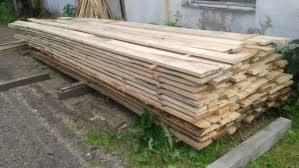 Stos surowej, jasnej tarcicy ułożony na drewnianych legarach na ziemi z fragmentami trawy i gruzu, przy szarej ścianie budynku.