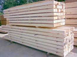 Ułożone w stosy deski z jasnego drewna, widoczne sęki i naturalna faktura, lekko rozmyte tło z zielenią drzew.