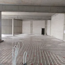 Rozległa podłoga w surowym stanie z betonowymi filarami i ścianami, pokryta systemem ogrzewania podłogowego z białymi rurkami ułożonymi w regularny wzór na specjalnej macie.