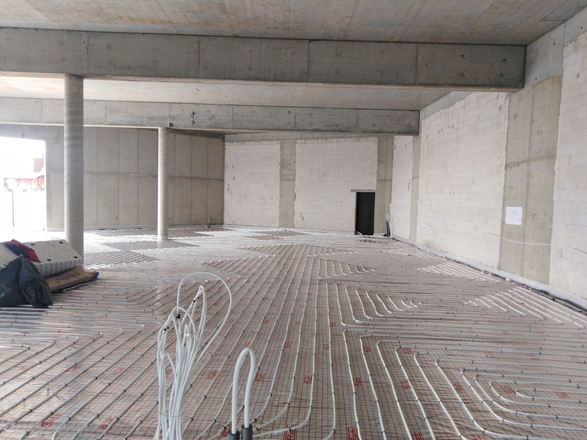 Rozległa podłoga w surowym stanie z betonowymi filarami i ścianami, pokryta systemem ogrzewania podłogowego z białymi rurkami ułożonymi w regularny wzór na specjalnej macie.