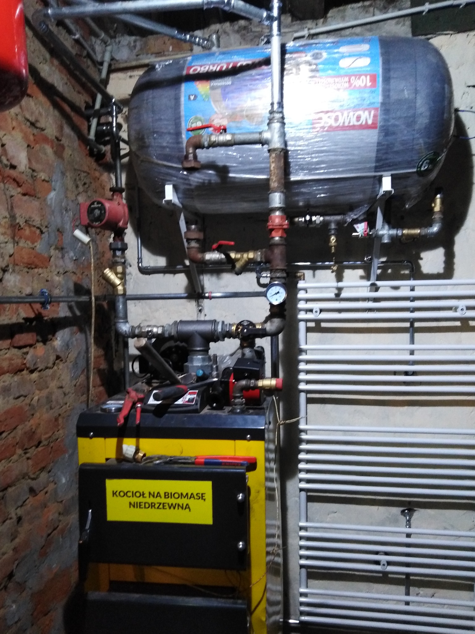 Kotłownia z kotłem na biomasę, zbiornikiem wody owiniętym folią i grzejnikiem drabinkowym, widoczne rury instalacji hydraulicznej i narzędzia.