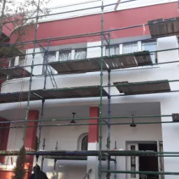 Budynek dwukondygnacyjny w trakcie malowania elewacji, widoczne rusztowania i częściowo pomalowana czerwono-biała fasada.