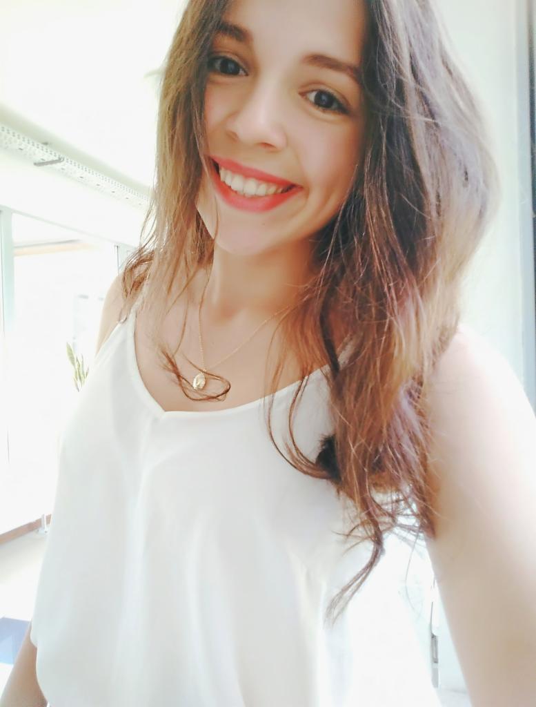 Uśmiechnięta młoda kobieta w białej bluzce na tle jasnego wnętrza z widocznym oknem. Widoczne włosy, naszyjnik i makijaż.