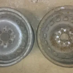 Dwie stalowe felgi samochodowe, jedna po prawej stronie z widocznymi śladami rdzy i starej farby, druga po lewej stronie po czyszczeniu, przygotowana do dalszej obróbki.