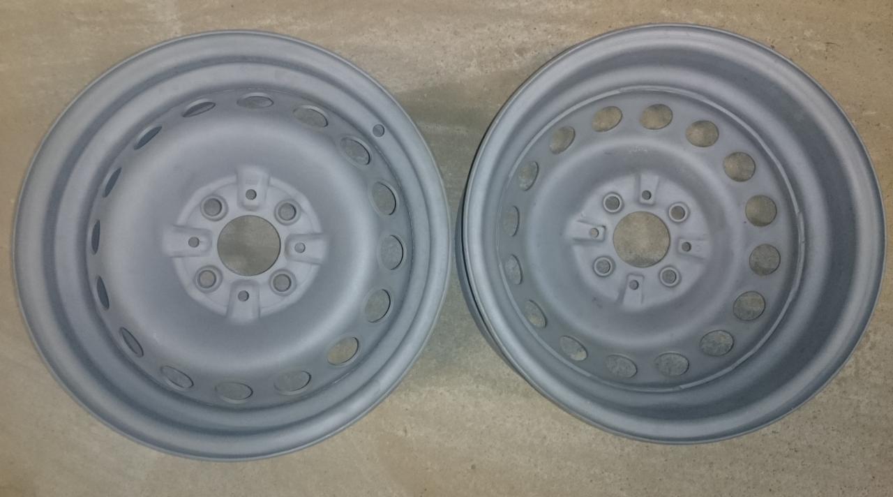 Dwie stalowe felgi samochodowe po obróbce, pokryte świeżą, jednolitą warstwą szarej farby, leżące na szorstkiej, betonowej powierzchni.