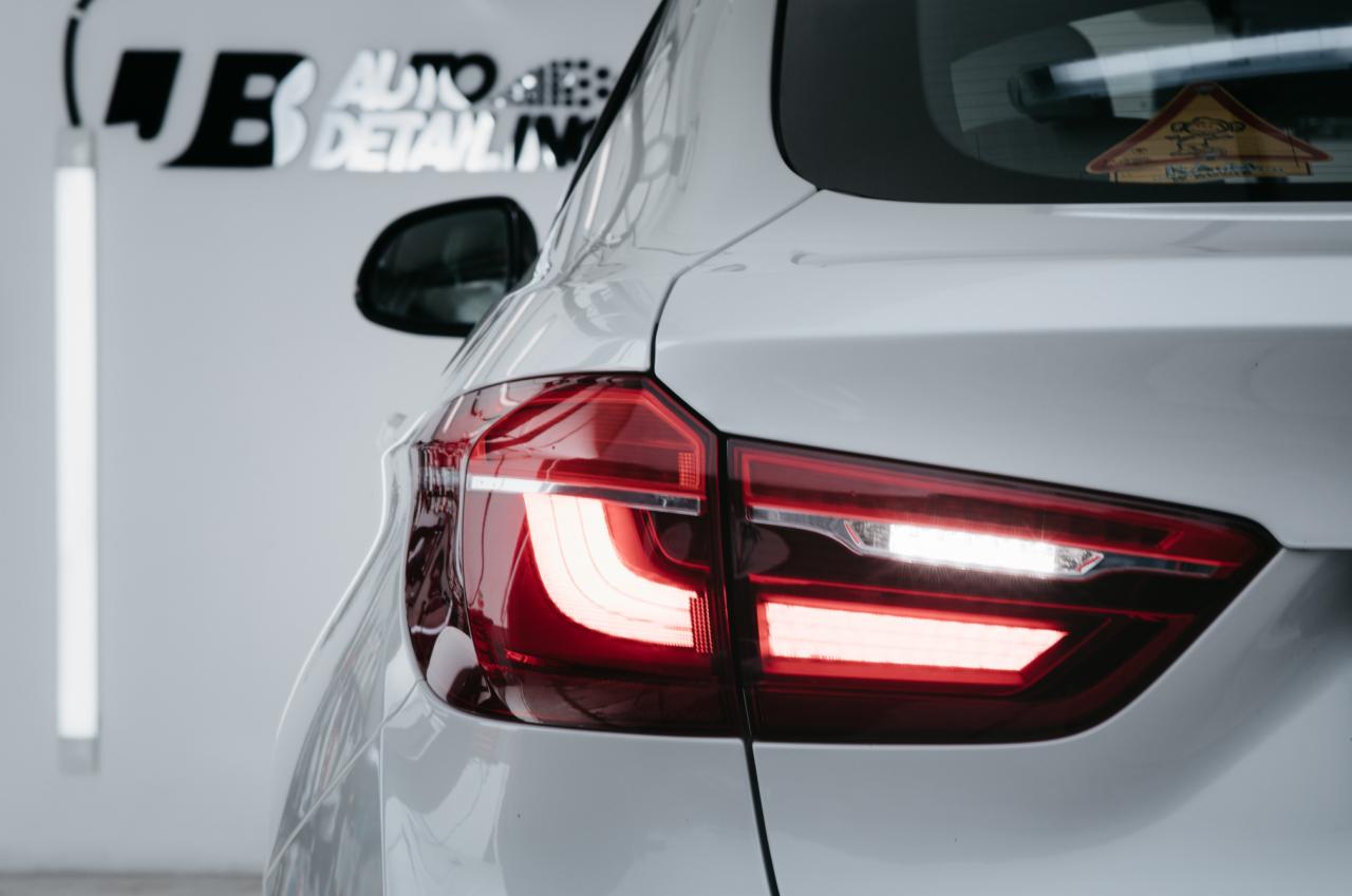 Tył białego samochodu z włączonymi światłami, widoczny znak ostrzegawczy dla dzieci na tylnej szybie i logo firmy detailingowej w tle.