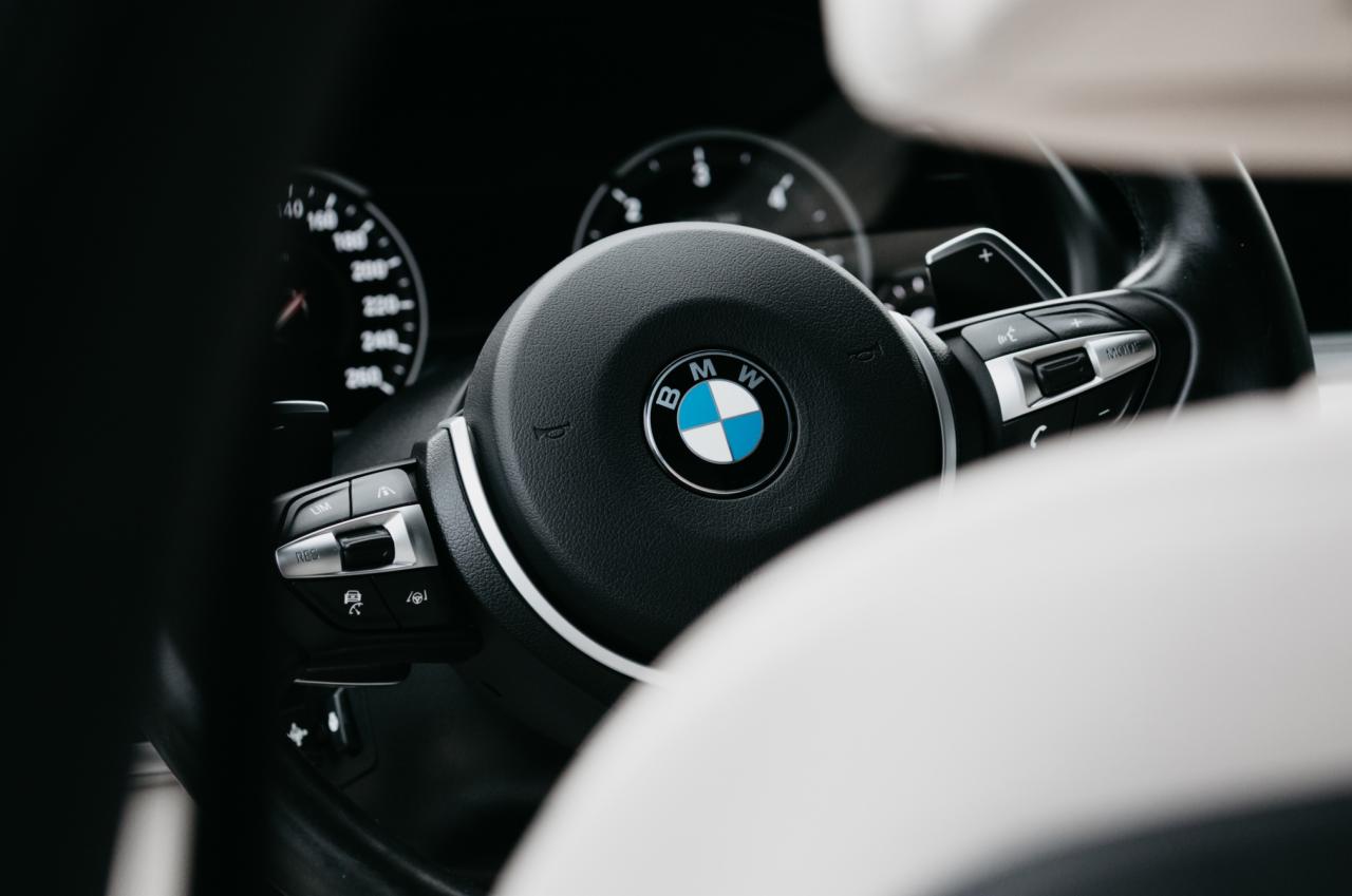 Wnętrze samochodu BMW, widok na kierownicę z logo, deskę rozdzielczą i fragment fotela w jasnej tapicerce.