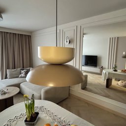 Usługi remontowo-budowlane - Jasne wnętrze salonu z beżową sofą, okrągłym stolikiem i designerską lampą wiszącą. W tle lustro powiększające przestrzeń i wpuszczające dodatkowe światło.