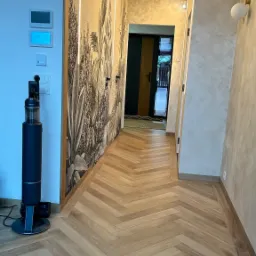 Beżowa podłoga ułożona w jodełkę w nowoczesnym korytarzu z oświetleniem LED i tapetą w roślinne motywy, widoczny odkurzacz pionowy.