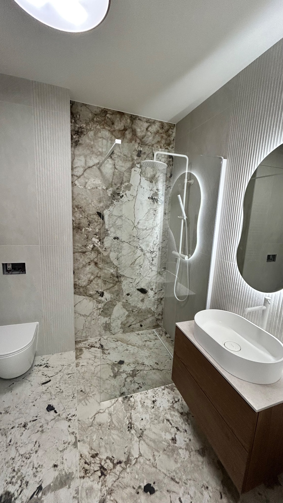 Nowoczesna łazienka z kabiną prysznicową typu walk-in, marmurowymi płytkami i podświetlanym lustrem. Umywalka nablatowa na drewnianej szafce. Minimalistyczny design.