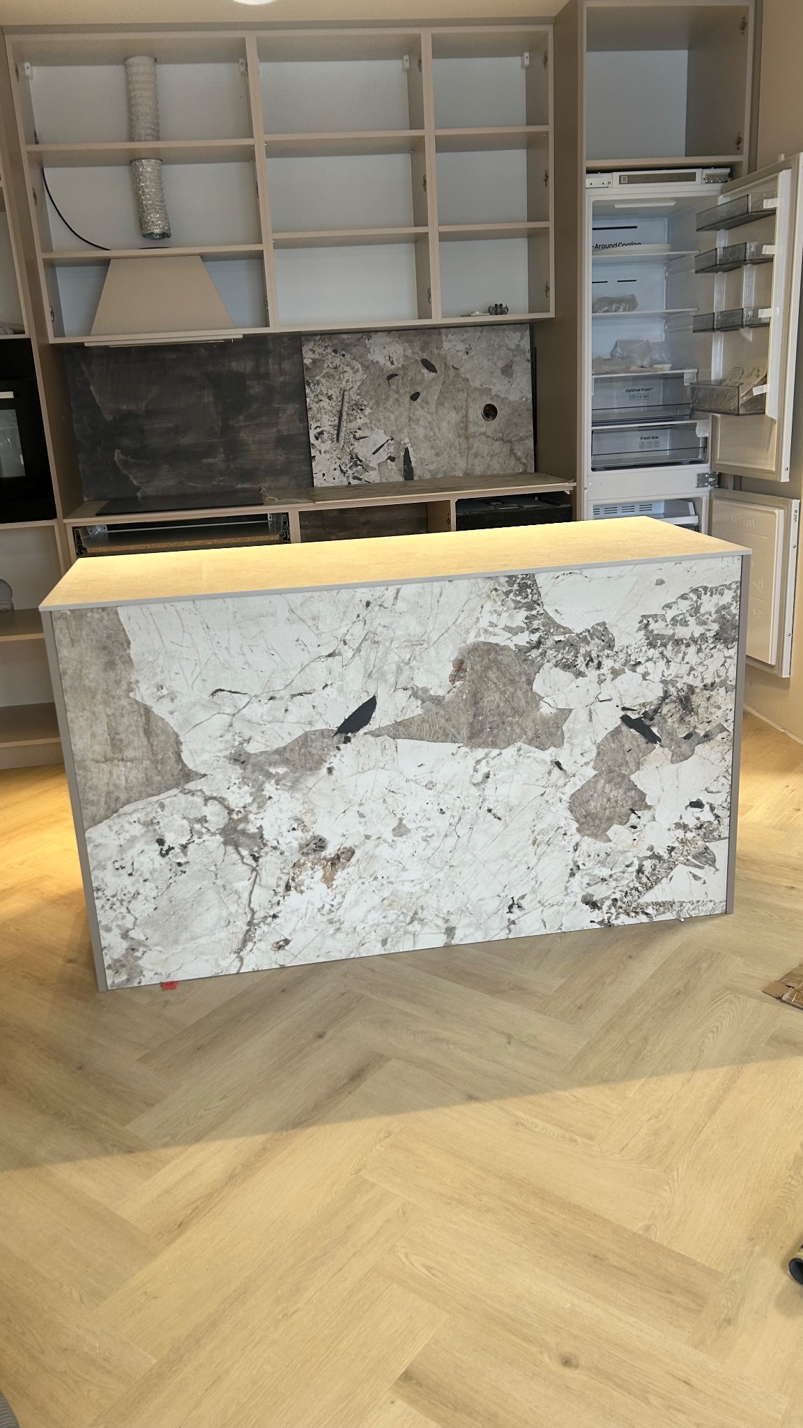 Wyspa kuchenna z blatem z marmuru w odcieniach bieli i szarości, na tle częściowo zamontowanych szafek kuchennych i otwartej lodówki. Parkiet ułożony w jodełkę.