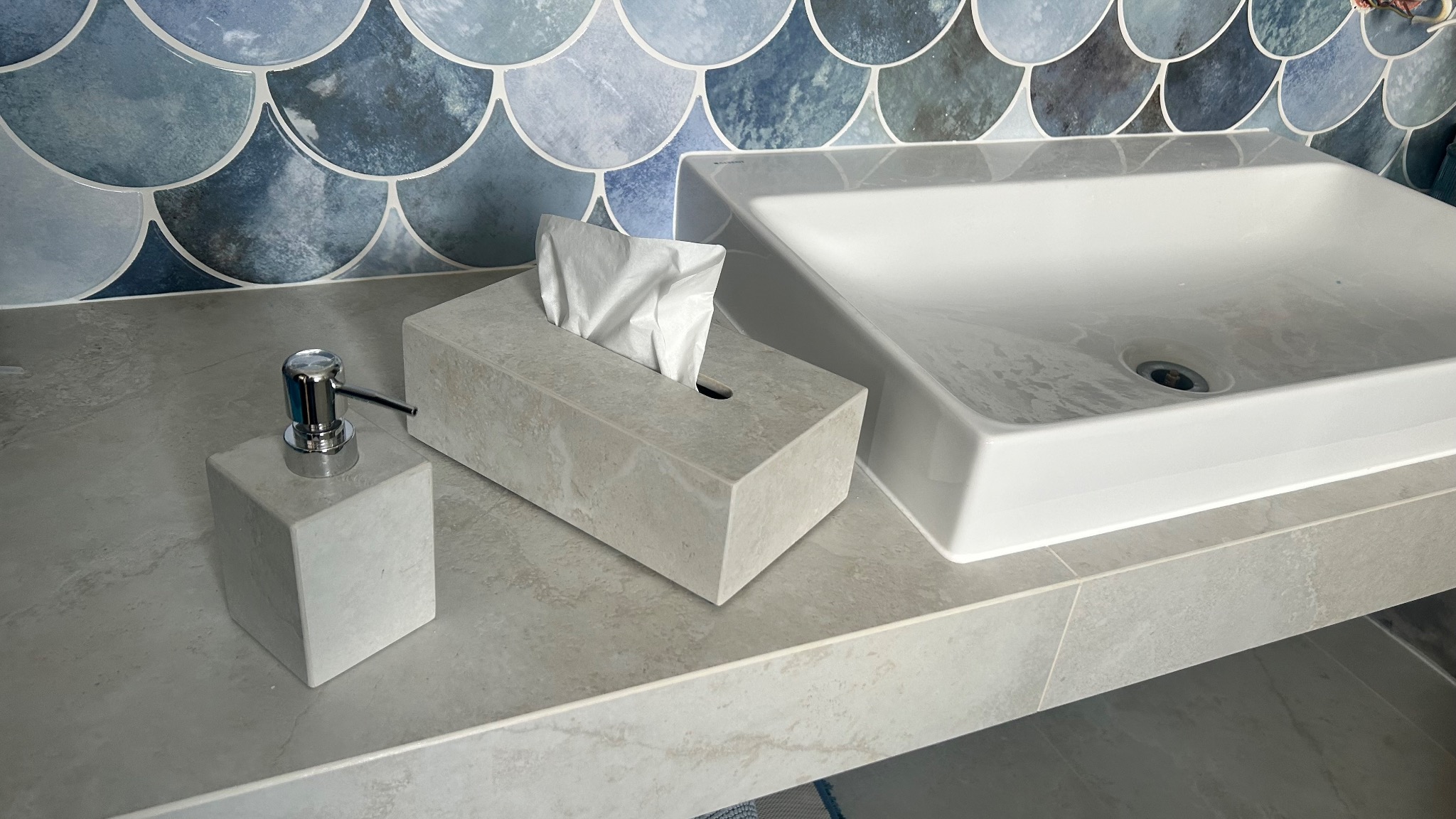 Biała, prostokątna umywalka nablatowa, dozownik mydła i pudełko na chusteczki z efektem marmuru na blacie z płytek. W tle niebieskie płytki w kształcie rybiej łuski.