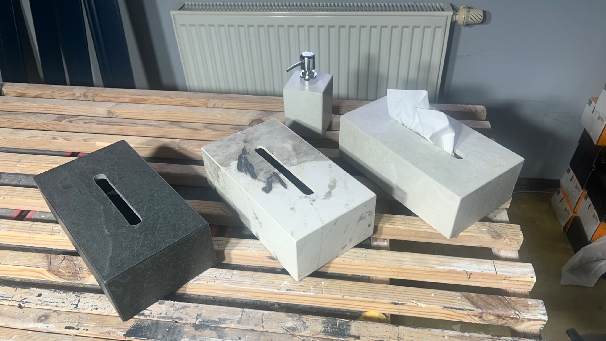 Betonowe akcesoria łazienkowe: dozownik mydła, pudełka na chusteczki, w odcieniach szarości i bieli, ustawione na drewnianej palecie, z grzejnikiem w tle.