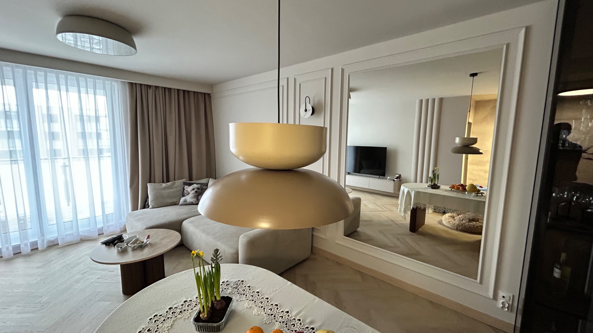 Jasne wnętrze salonu z beżową sofą, okrągłym stolikiem i designerską lampą wiszącą. W tle lustro powiększające przestrzeń i wpuszczające dodatkowe światło.