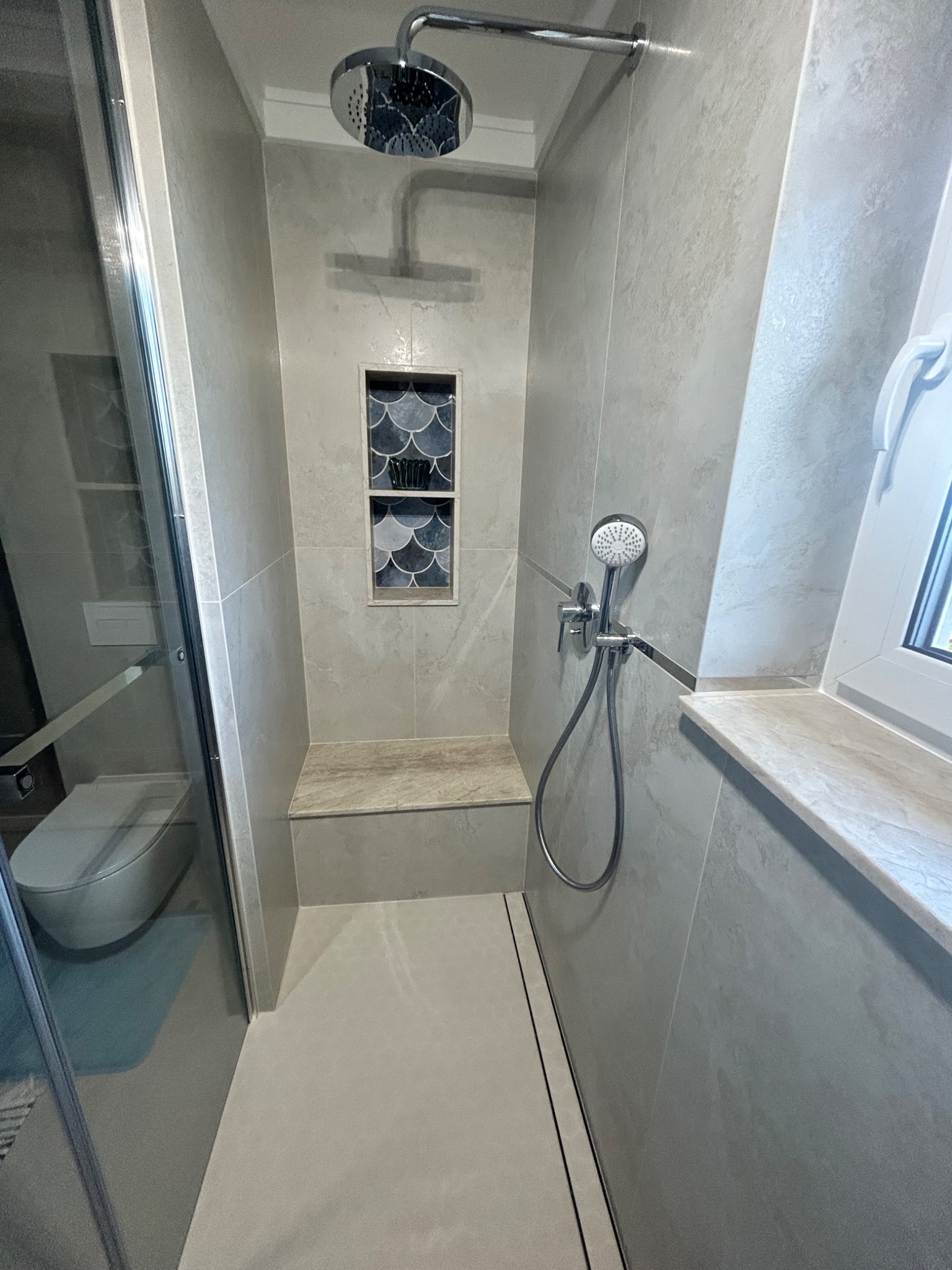 Nowoczesna kabina prysznicowa z siedziskiem, wykończona płytkami w odcieniach szarości. W ścianie wnęka z dekoracyjną mozaiką w kształcie rybiej łuski.