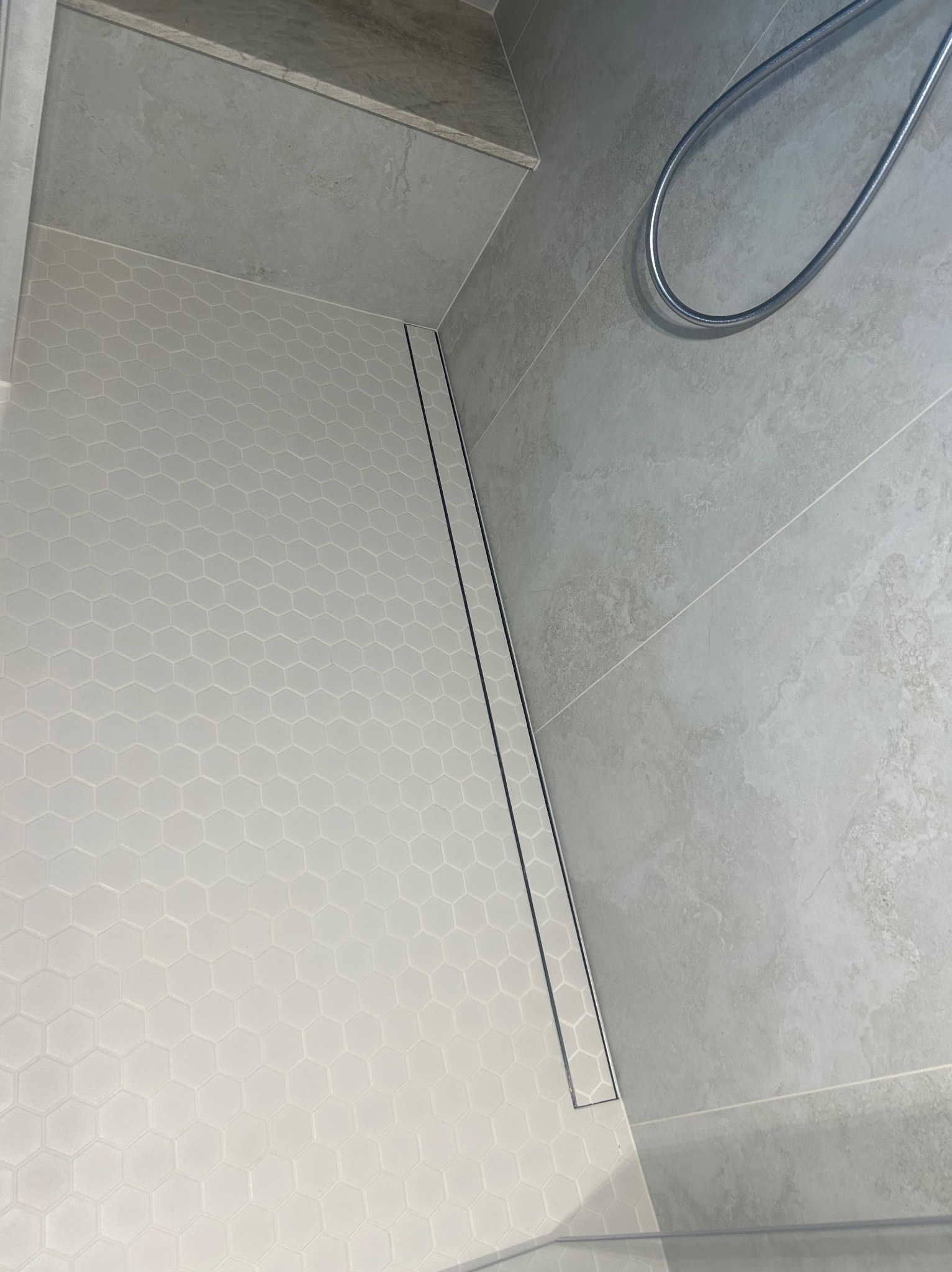 Jasna łazienka z heksagonalną mozaiką podłogową i odpływem liniowym. Minimalistyczny design, szare płytki ścienne i chromowana słuchawka prysznicowa.